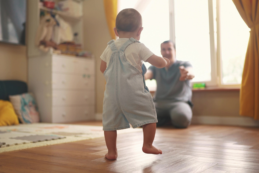 Kleinkind in Jeansoverall läuft auf einen Erwachsenen zu, der mit offenen Armen in einem hellen Raum steht.